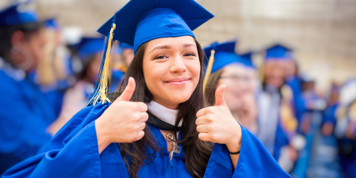 Young college girl in cap and gown at the graduation ceremony giving the thumbs up for success.
