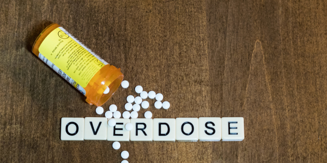 Overdose spelled with tile letters in a row with small white pills and an open prescription bottle. Photographed from above on a wooden table. Image has copy space.
